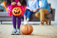 Child in costume holding a pumpkin basket with parents sitting on couch in background